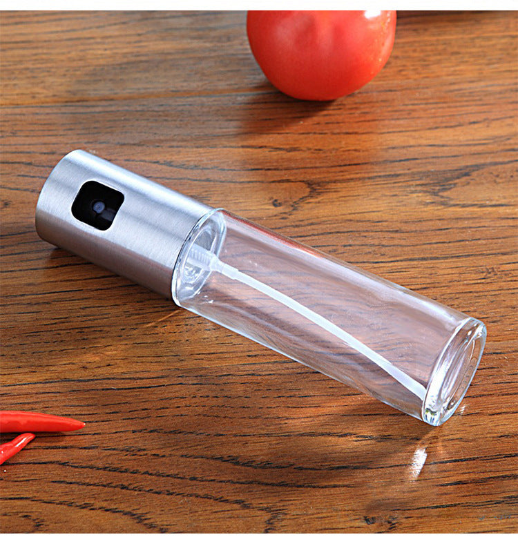 Stainless steel oil sprayer with clear glass bottle for kitchen cooking, shown on wood table with tomato